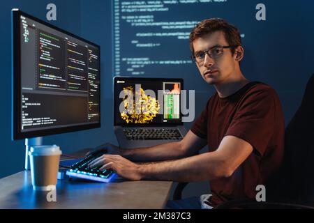 Portrait of Intelligent male programmer working on pc writing brand new code at his home office, looking at camera on background with digital wall with application info page. Data science concept Stock Photo