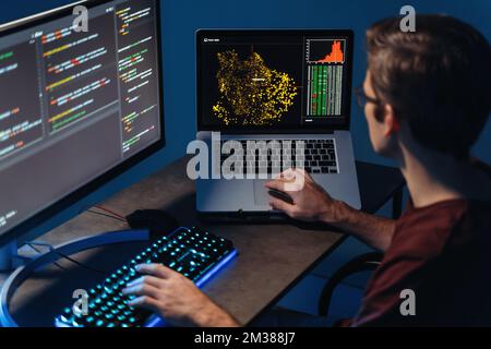 Back view of busy male data scientist typing on keyboard on pc and laptop with programming code on screen, using php language for coding app, sitting at home office Stock Photo