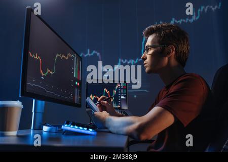 Side view of young man trader analyst at working table, analyzing financial chart, stock quotes, share prices, trading online, checking data on cryptocurrency graph on computer screen, making notes Stock Photo