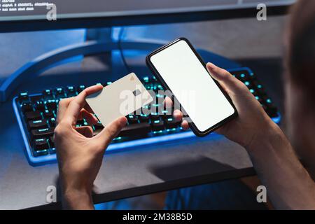 Close-up of male hands holding credit card and smartphone with white mockup copy space screen for your advertising content. Gamer buying access to new level using phone and bank card.  Stock Photo