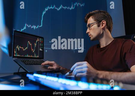 Financial crypto expert in glasses working late night online on laptop at home office, analyzing candlestick chart looking at screen of monitor, on digital wall with diagram Stock Photo