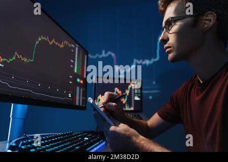 Closeup of intelligent crypto trader inventing successful trading strategy, reading candlestick chart, trying to predict trends on trading market, writing in notepad, looking at pc monitor at night Stock Photo