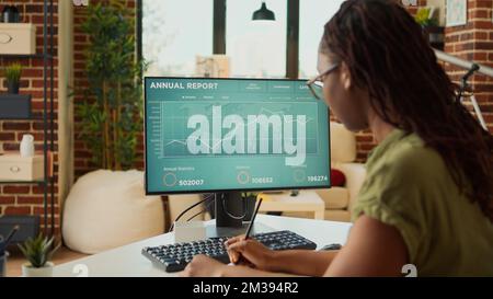 Young adult looking at annual sales report data on computer, analyzing accounting statistics on web. Woman working remote with financial diagrams, marketing education at home. Stock Photo
