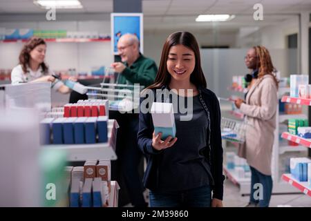 Woman customer looking at shelves with pharmaceutical products, choosing to buy vitamin supplements for for immune system. Pharmacy full with clients and health care medication. Stock Photo