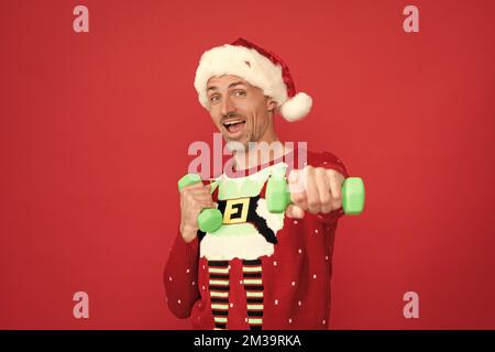 Happy guy in Christmas hat and jumper do dumbell exercises red background, Santa Stock Photo