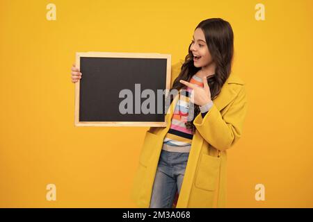 Teenager younf school girl holding school empty blackboard isolated on yellow background. Portrait of a teen female student. Happy teenager, positive Stock Photo