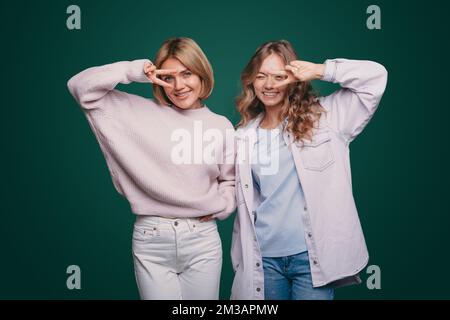 Photo of pretty two women showing v-sign symbols looking at camera isolated over green color background. Number two. Stock Photo