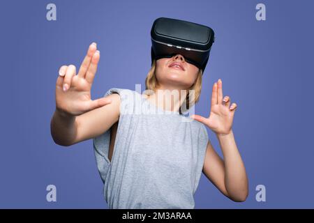 Woman getting experience using VR-headset glasses of virtual reality, gesticulating with hands isolated over blue background. Digital technology Stock Photo