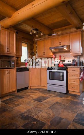 Wooden shaker style cabinets and modern appliances in kitchen with ceramic tile floor inside log cabin. Stock Photo