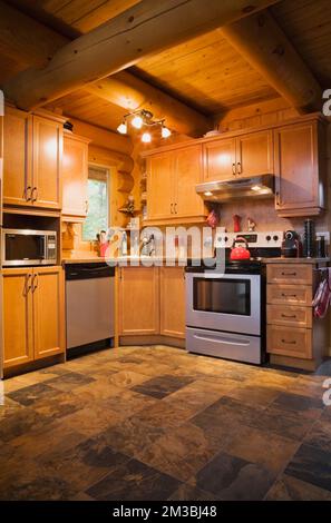 Wooden shaker style cabinets and modern appliances in kitchen with ceramic tile floor inside log cabin. Stock Photo
