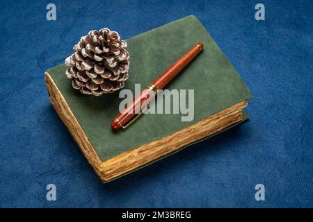 retro leather-bound journal with decked edge handmade paper pages, decorative frosty pine cone and a stylish pen, journaling and winter holidays conce Stock Photo