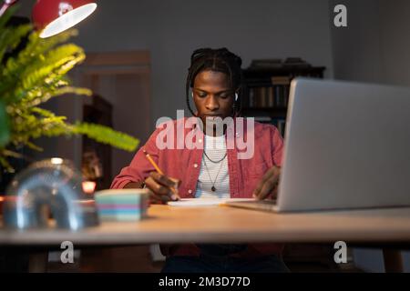 Hardworking young African guy student writing notes while watching webinar on laptop at home Stock Photo