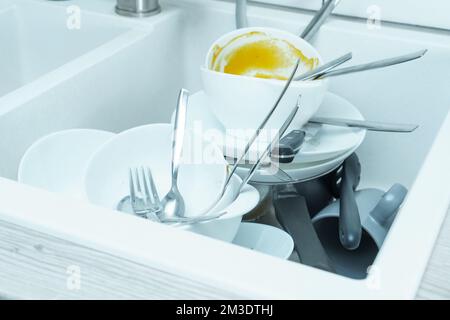 Close-up of white sink full of dirty white tableware, ceramic plates, bowls, knives, forks, spoons and grey cups. Cooking, housekeeping, cleaning, ute Stock Photo