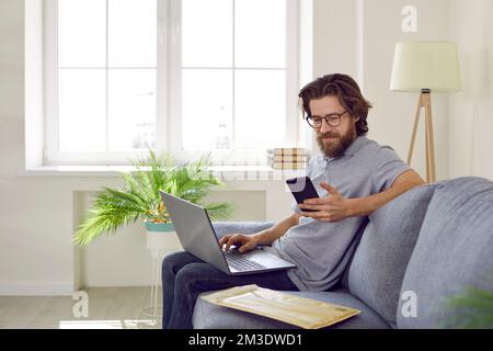 Happy man sitting on comfortable couch at home, using mobile phone and working on laptop Stock Photo