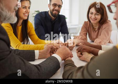 Team of happy supportive business people sitting together at office table and holding hands Stock Photo