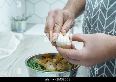 Cropped photo of man wearing grey apron peeling hardboiled egg over metal bowl full of carrot, onion peelings, standing at grey wooden table in kitche Stock Photo