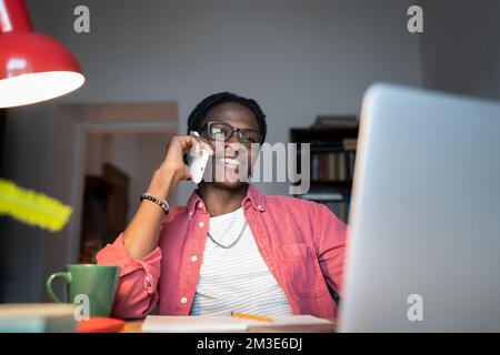 Friendly young African man freelance consultant talking to client on phone while working from home Stock Photo