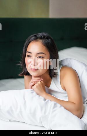 Vertical shot of beautiful asian woman lying in her bed on pillow, covered in duvet, looking thoughtful and smiling, waking up and thinking Stock Photo