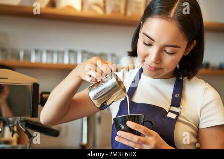 Smiling asian barista girl, making coffee, pouring steamed milk into cappuccino, doing latte art in cup, working in cafe Stock Photo