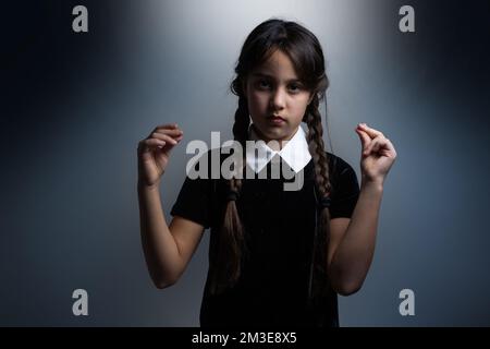 Portrait of little girl with Wednesday Addams costume during Halloween. Serious expression and dark atmosphere with dark background. Stock Photo