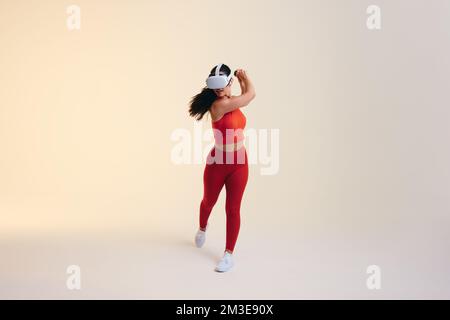 Sporty young woman playing tennis in virtual reality. Athletic young woman swinging a virtual tennis bat using gaming controllers. Sportswoman wearing Stock Photo