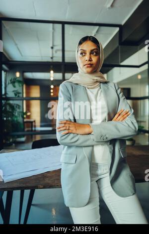 Ethnic Muslim businesswoman looking away thoughtfully while standing in an office boardroom. Young female entrepreneur wearing a hijab in a modern wor Stock Photo