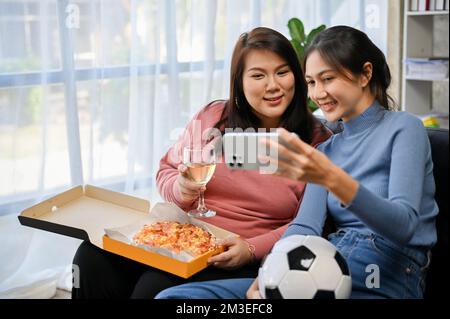 Two happy millennial Asian female friends sit on the sofa and taking selfie with a smartphone while watching a football match and having pizza party t Stock Photo