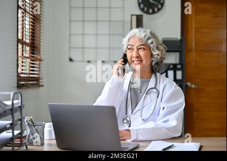 Happy and professional Asian aged female doctor talking on the phone with someone while working in her office. Stock Photo