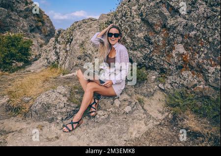 Young woman freelancer traveler working online anywhere outdoors using laptop enjoying mountain peak view. Happy female downshifter in sunglasses holding computer in unusual workplace at summer Stock Photo
