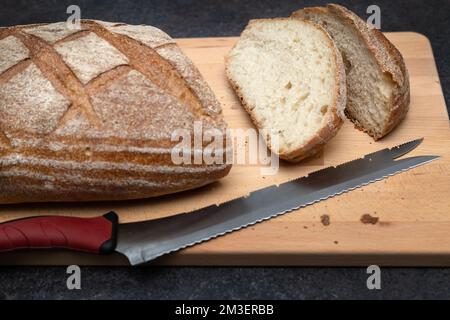 Oval loaf of freshly baked sourdough slided bread with knife on cutting wooden board, angle view. Rustic sourdough bread on dark gray table Stock Photo