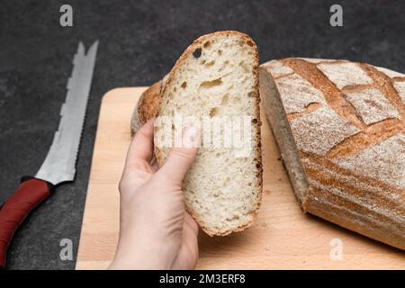 holding slice of oval freshly baked sourdough bread in hand on cutting wooden board with knife, angle view. Rustic sourdough bread on dark gray table Stock Photo