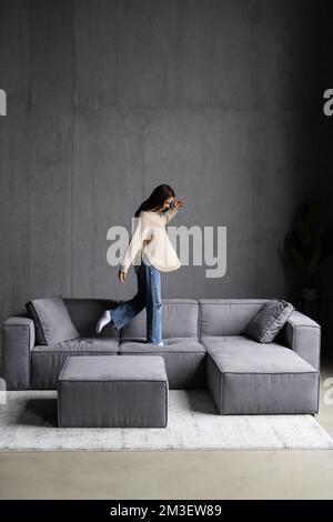 Full body beautiful woman meloman in casual clothes and wireless headphones raising arms, jumping and dancing near sofa while having fun in living roo Stock Photo