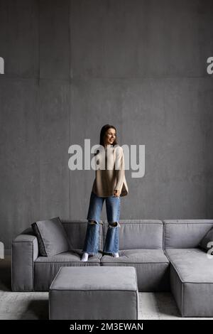 Full body beautiful woman meloman in casual clothes and wireless headphones raising arms, jumping and dancing near sofa while having fun in living roo Stock Photo