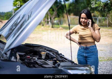 Young Asian woman using a mobile phone to call the mechanic while looking at broken down car on street. Stock Photo