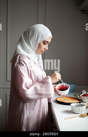 Side view portrait Muslim woman cooks dessert cake at kitchen, arabian young model in hijab and abaya. Islamic traditional clothing Stock Photo