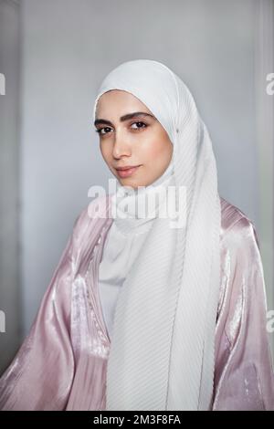 Portrait of young Muslim woman in white hijab headscarf and light pink abaya on wall background. Arab beautiful model Stock Photo