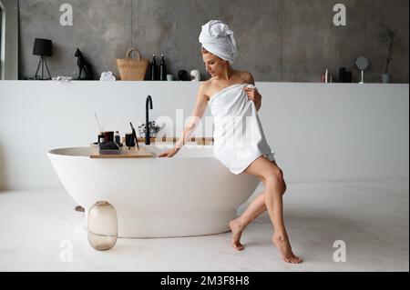 Calm woman wrapped in white cotton towel standing near bath in bright hotel room Stock Photo