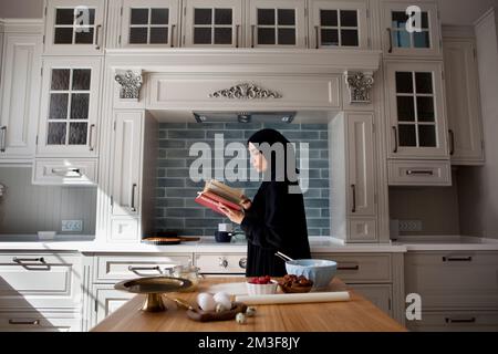 Beautiful Muslim woman cooks in kitchen and reads book, wears black abaya and hijab Stock Photo