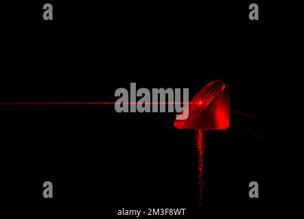 A red laser beam striking a prism. The beam was made visible by a fine spray of water. Water drops on prism, and water streaks in beam. Stock Photo
