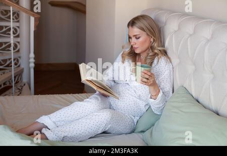 Pregnant woman sitting at home in bed, reading book and drinking tea or water from cup Stock Photo