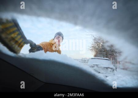 Man clearing snow from windshield his snowy car after snowfall. Parking on city street in winter. Stock Photo