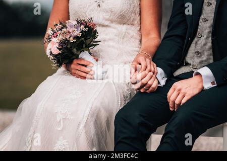 A closeup shot of a newly-wed couple sitting on a bench holding hands with each other Stock Photo