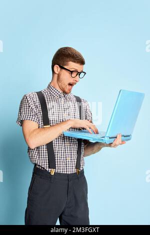Young man in checkered shirt and suspenders emotionally typing on laptop isolated over blue background. Program developer Stock Photo