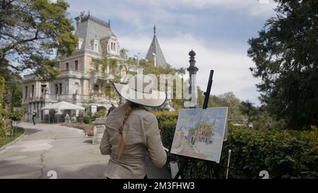 The painter paints a picture of the fortress. Action. Rear view of an elegant woman artist in a hat painting a castle and green vegetation Stock Photo