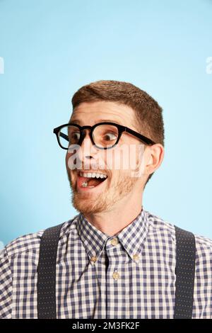 Portrait of young man in checkered shirt, shorts and suspenders with laptop isolated over blue background. Extremely excited. Good news Stock Photo
