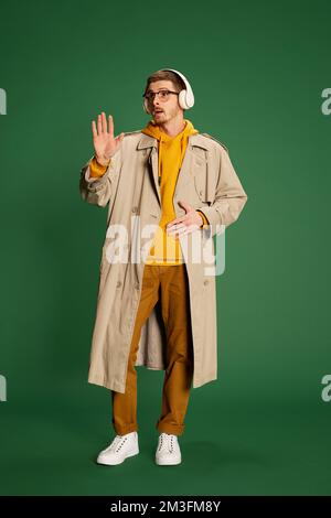 Portrait of young stylish man in trendy clothes posing in headphones isolated over green background. Attention Stock Photo