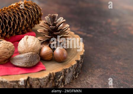 Festive still life with space for copy Christmas holidays post containing nuts and pinecones on a wooden block with red napkin and dark background. Stock Photo