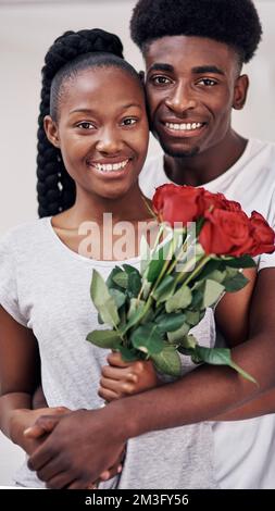 She deserves flowers on all days of the week. a young woman holding a bunch of red roses while standing with her boyfriend at home. Stock Photo