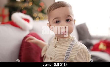 Adorable toddler standing by christmas tree with serious expression at home Stock Photo