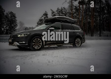 12-12-2022 Riga, Latvia  a black car with a surfboard on top of it in the snow with trees in the background and a fence. . Stock Photo